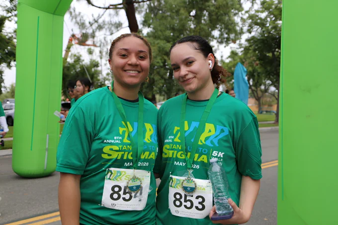 Two individuals wearing green race shirts and medals, standing under a green inflatable arch. They have race bibs numbered 854 and 853, participating in a 5K race. One is holding a bottle of water and both are smiling at the camera.