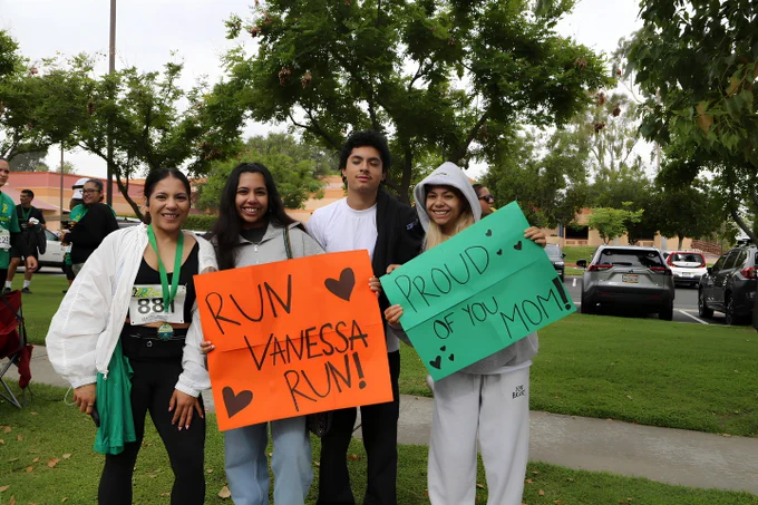 A group of individuals standing outdoors, holding colorful signs. One sign reads "Run Vanessa Run" with hearts, and the other says "Proud of You Mom!" They are on a grassy area with parked cars and trees in the background.