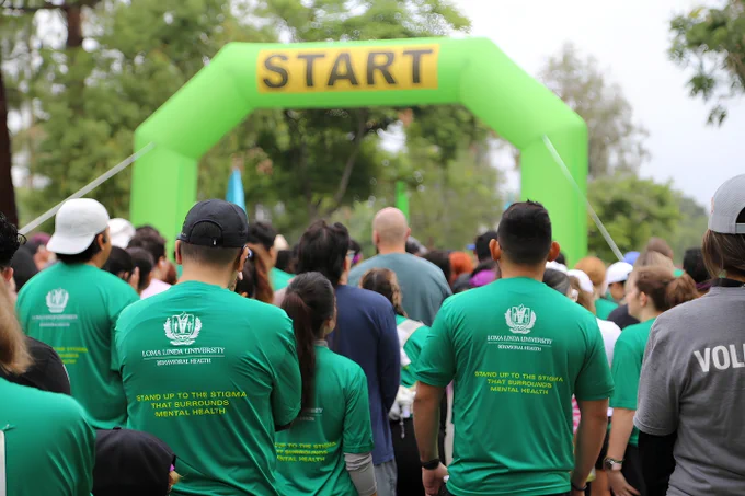 A group of participants wearing green shirts with "Loma Linda University" logos are gathered at the starting line of a race, indicated by a large green inflatable arch marked "START." Trees and other participants are visible in the background.