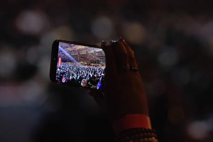 A person holds a smartphone capturing a crowded concert hall with bright stage lights. The phone's screen shows a clear view of the audience inside the venue.