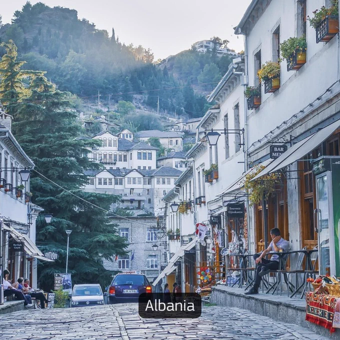 Image of a street in Albania with stone-paved roads, small shops and cafés, and hillside buildings in the background.