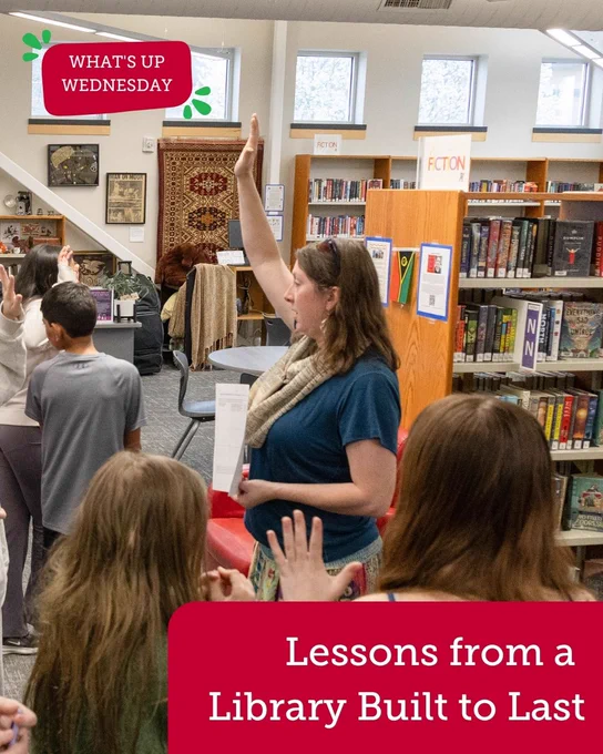 A person raises a hand while standing among a group of individuals inside a library. Bookshelves filled with books are in the background. A sign reads "Lessons from a Library Built to Last.