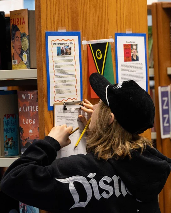 A person wearing a Disney sweatshirt and a cap is writing with a pencil on a clipboard attached to a wooden shelf in a library. The shelf displays books including "With a Little Luck," "Aristotle and Dante," and "Furia." There are informational sheets on the shelf, one with a flag, and one with text and an image.