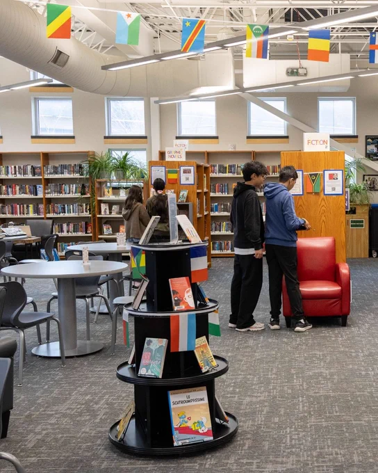 A library interior featuring a variety of bookshelves and a red armchair. Flags are displayed above. Several individuals are browsing books near a shelf labeled "Fiction." In the foreground, a round display shelf holds books with colorful covers.