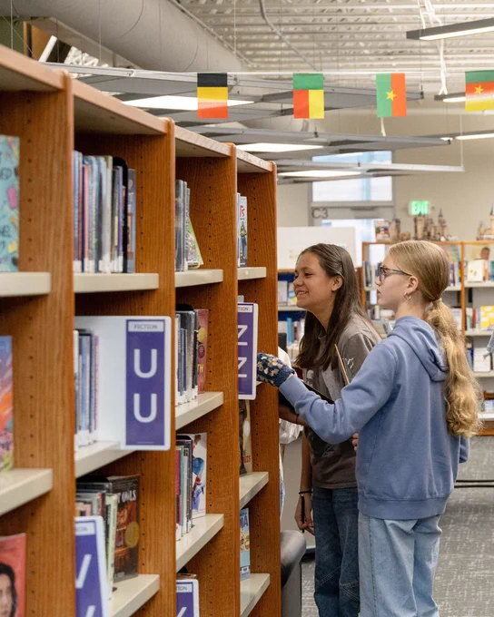 Two people are standing in a library, looking at books on a shelf. The shelves are organized alphabetically. Small flags hang from the ceiling in the background.