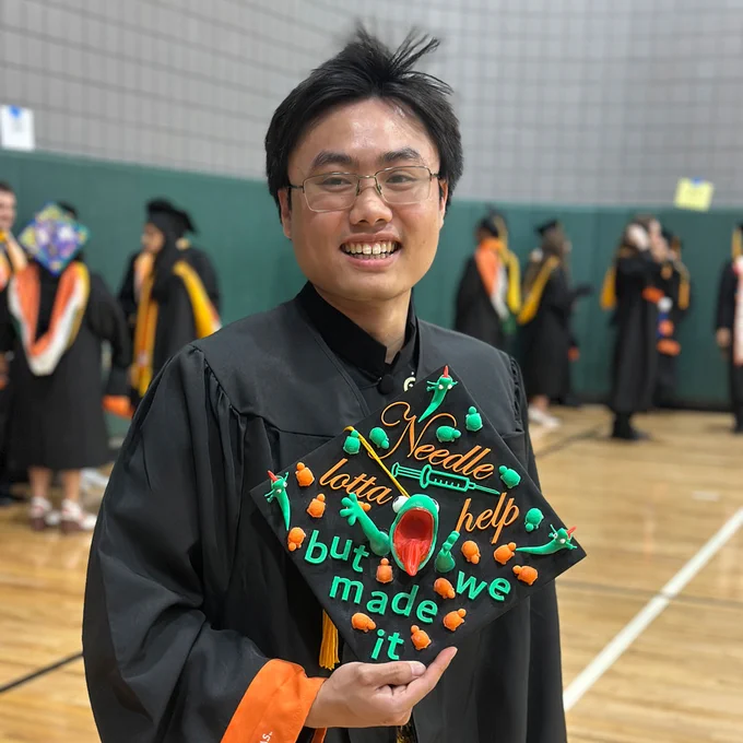 A student in a graduation gown is smiling and holding a decorated graduation cap. The cap features colorful designs with the words "Needle lotta help but we made it" and images of syringes and caps. Other graduates are visible in the background.