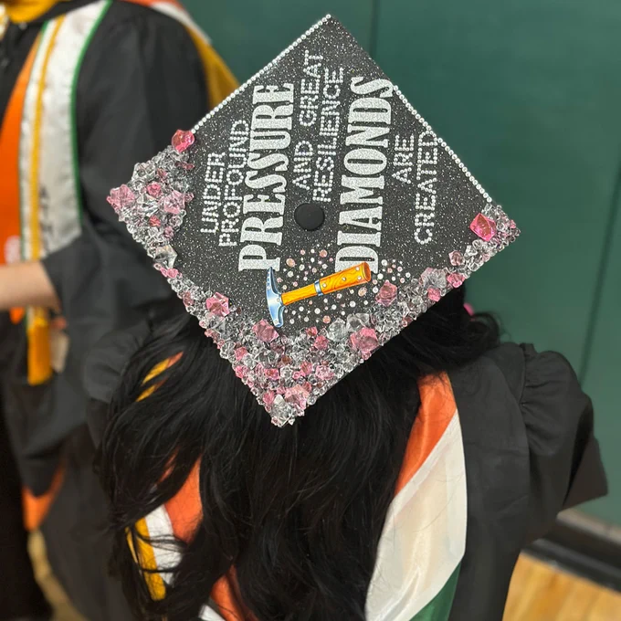 A graduate is wearing a decorated mortarboard cap. The cap is adorned with shiny stones and displays the phrase "Under profound pressure and great resilience, diamonds are created" along with an illustration of a hammer.