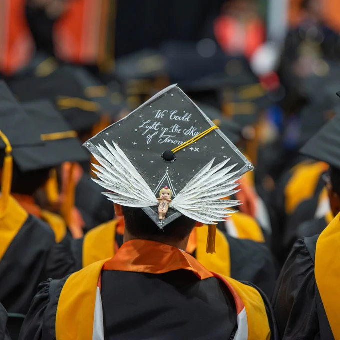 A graduate wearing a decorated cap with a figure holding wings, surrounded by students in graduation gowns. The cap includes the phrase "We Could Light up the Sky of Tomorrow".