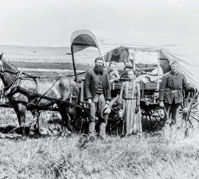 A family of four stands beside a covered wagon hitched to two horses in an open prairie landscape.
