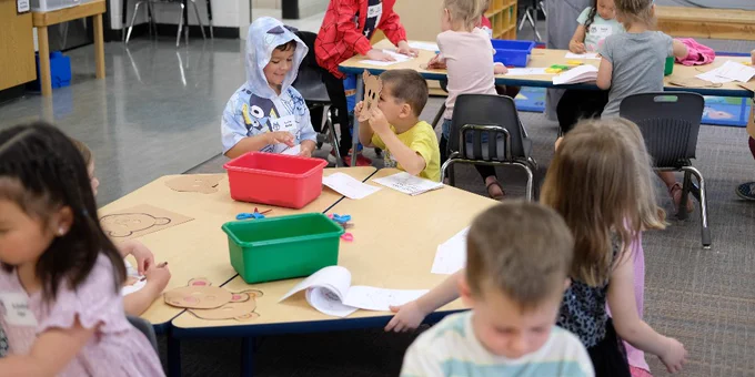 Young children engaged in various activities at classroom tables with a teacher standing nearby.
