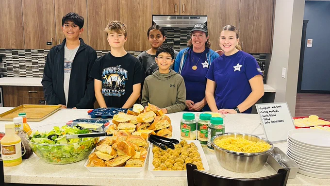A group of five people standing behind a table filled with sandwiches, salad, pasta, and condiments in a kitchen setting.