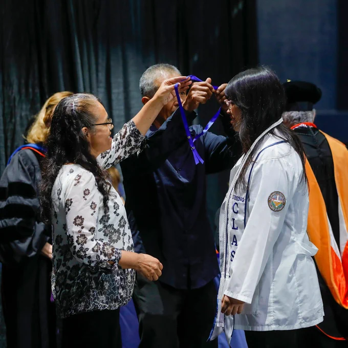 Two individuals are in the process of placing a ceremonial ribbon over the shoulders of a graduate from the UTEP College of Nursing who is wearing a white coat with the word "CLASS" visible. There are members of the college's faculty in academic regalia in the background.
