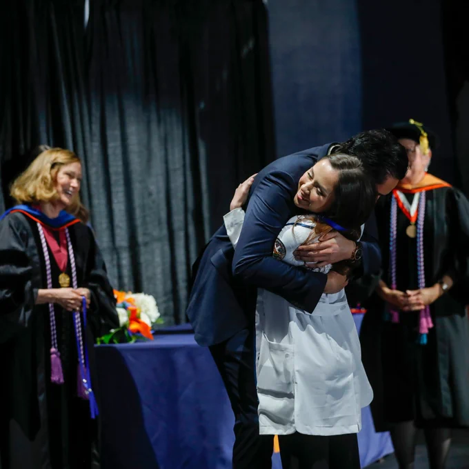 A heartwarming moment at a Pinning Ceremony for the UTEP College of Nursing, where a man and a woman embrace on stage. They are surrounded by several members of the college's faculty in academic regalia, and there is a table with flowers in the background.