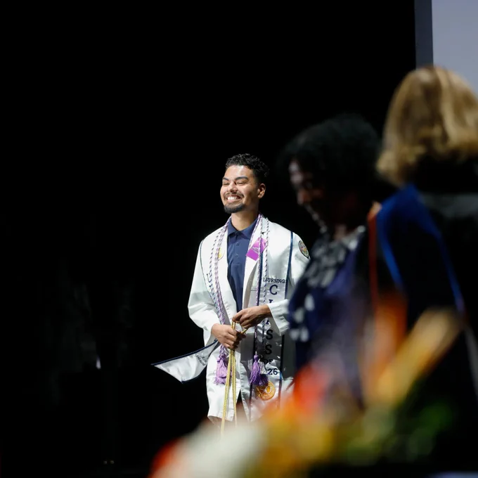 A graduating student from the UTEP College of Nursing stands on a stage, during the college's pinning ceremony, wearing a white coat adorned with sashes and a stethoscope, smiling. The foreground shows blurred figures and vibrant flowers.