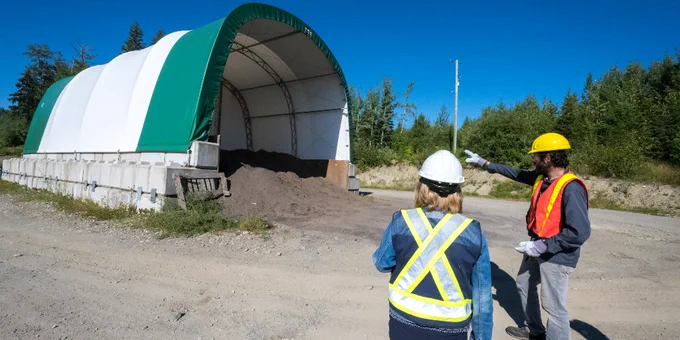 A person gestures toward a storage building containing a pile of dark material. Another individual wearing a safety vest and helmet observes. The scene is set outdoors on a clear day, surrounded by greenery.