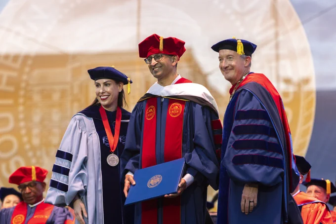 Three individuals in academic regalia are on a stage at a graduation ceremony. One person in the middle is holding a diploma folder. In the background is a large seal for SMU.