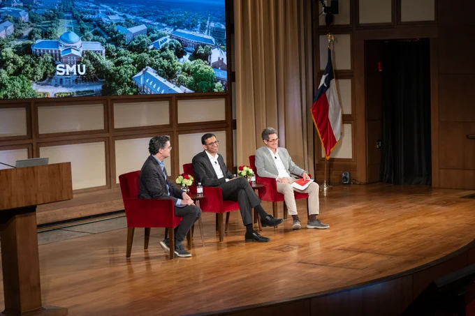 Three individuals are seated on a stage with red chairs, engaged in a panel discussion. A screen behind them displays an aerial view labeled "SMU." A Texas flag is visible in the background.