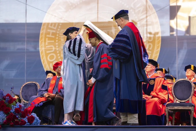 A university graduation ceremony where a graduate is being hooded by two officiants in academic regalia, with a large emblem in the background and others seated on stage.