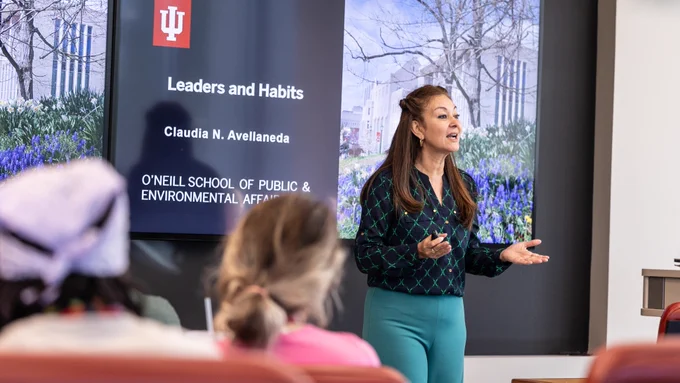 Professor Claudia Avellaneda is speaking in front of a classroom of students. The presentation screen behind her displays the Indiana University logo and the title "Leaders and Habits." 