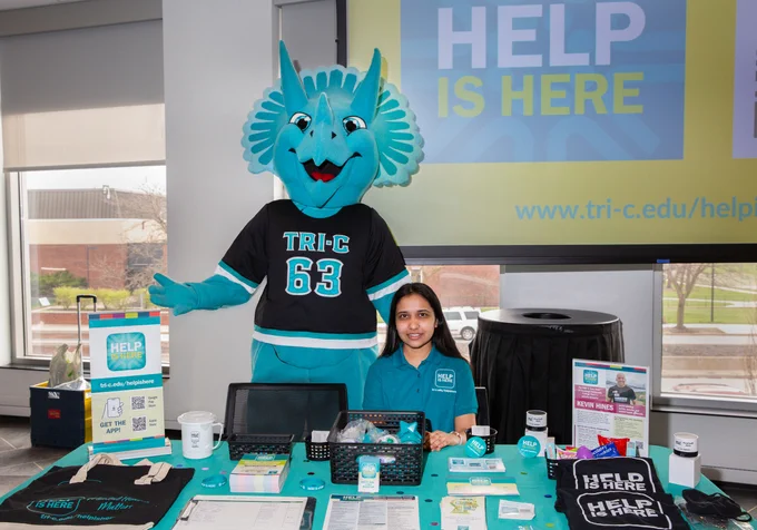 A dinosaur mascot in front of a sign that says "Help is Here"