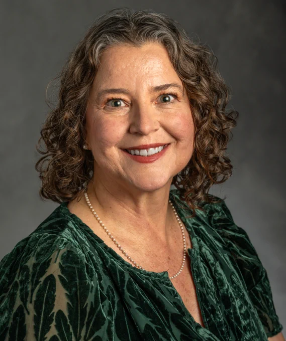 Mary Juzwik headshot. Juzwik wears a green velvet blouse and a pearl necklace. Juzwik's hair is dark and curly and falls about shoulder-length. 
