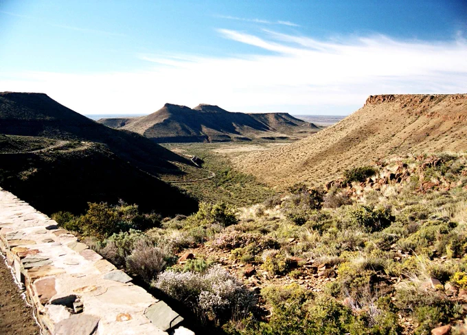 A panoramic view from a high vantage point overlooking the Karoo National Park in South Africa. A winding road snakes through a vast valley surrounded by arid, flat-topped mountains under a bright blue sky with wispy clouds.