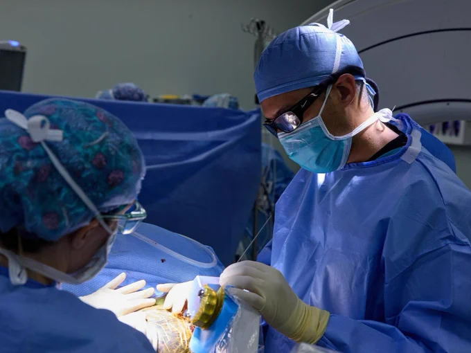 Medical professionals performing a LITT (Laser Interstitial Thermal Therapy) procedure in an operating room, wearing blue scrubs, masks, and caps. One person is focused on the procedure, while the other assists. The environment includes typical surgical equipment.