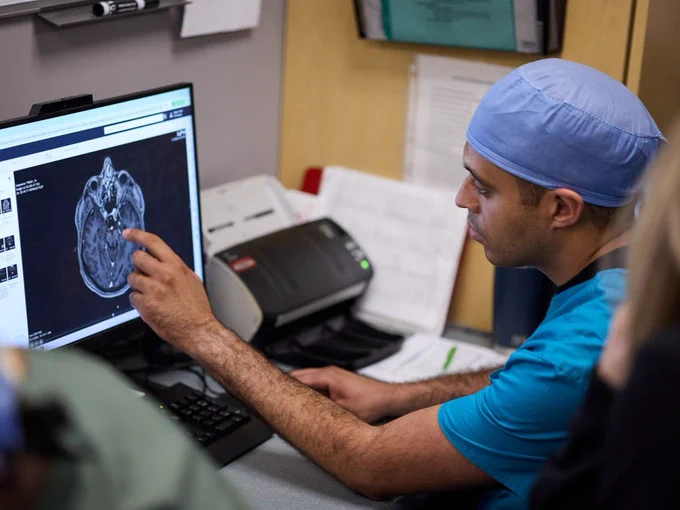 A person in a medical uniform and cap is seated at a desk, pointing at a brain scan displayed on a computer monitor. The setting appears to be a medical office or hospital.