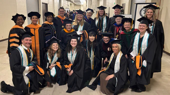 A group of people in graduation gowns and caps stand in a hallway, smiling. The attire includes various academic regalia, with some wearing orange and black robes. In the background, more people in similar attire are visible.