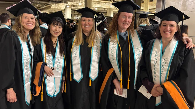 A group of five people wearing graduation caps and gowns stand together, smiling. They are all wearing sashes with "Cohort Inaugural Class" written on them. The setting appears to be indoors, possibly at a graduation ceremony venue.