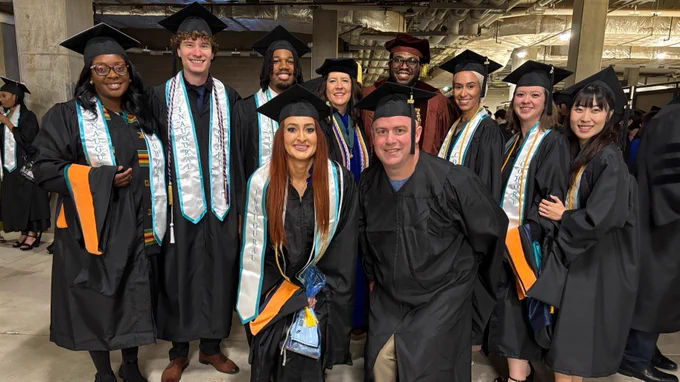 A group of graduates wearing caps and gowns, some with colorful stoles, standing together in an indoor setting. They all appear to be smiling and celebrating.