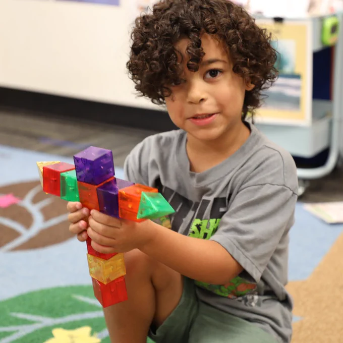 A child with curly hair is sitting on a colorful carpet, holding and showing a structure made of magnetic tiles in various colors. Shelves with educational materials are visible in the background.
