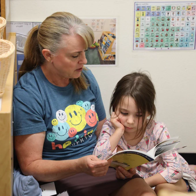A person sits on the floor with a child, reading a book together. The person is wearing a shirt with colorful smiley faces. Educational posters are on the wall.