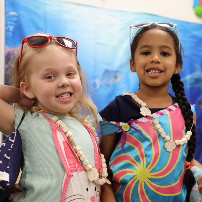 Two children smiling at the camera wearing colorful outfits and seashell necklaces. One child is wearing red sunglasses, and the other has light blue sunglasses. There is an ocean-themed backdrop with illustrations of marine life.