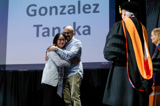 Two people sharing a hug on stage during a pinning ceremony, with a sign displaying the names 'Gonzalez' and 'Tanaka' in the background. An official in academic regalia stands nearby, facing them.