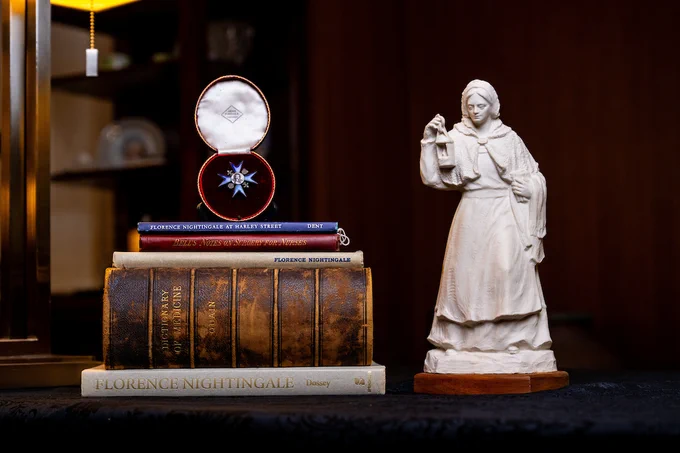 A marble statue of Florence Nightingale holding a lamp stands on a table. Beside it, there is a stack of books, with titles including "Florence Nightingale" and "The Memoirs of Florence Nightingale." On top, there is an open display case containing a decorative medal.