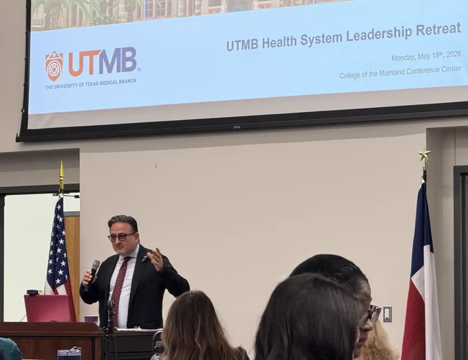 dr. Jochen Reiser stands at a podium during the UTMB Health System Leadership Retreat. An American flag is positioned nearby. The screen above displays the UTMB logo and event details, including "College of the Mainland Conference Center" and the date, May 18th, 2026. Several attendees are seated in the foreground.