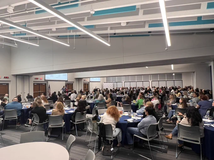 A large conference room filled with many people seated at round tables, attentively facing a presentation area. The room has modern lighting and a high ceiling. American flags are positioned near the exits.