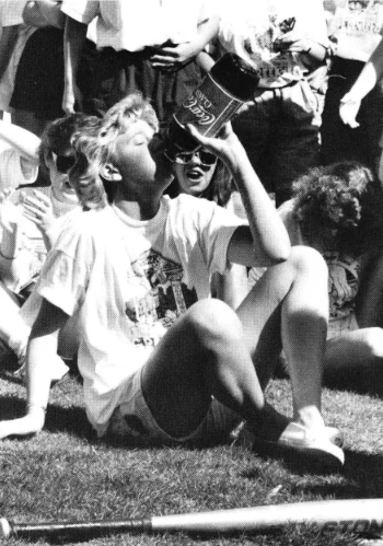 A person sitting on grass is drinking from a large Coca-Cola bottle. They are surrounded by several other people who appear to be enjoying an outdoor gathering.