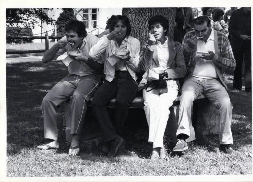 Four people are sitting on a bench outdoors, eating from bowls. The background features trees and grass, indicating a park or garden setting.