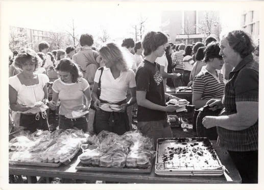 A large group of people gathered around tables outdoors, selecting items from trays of sandwiches and baked goods. The setting appears to be a casual event or gathering.