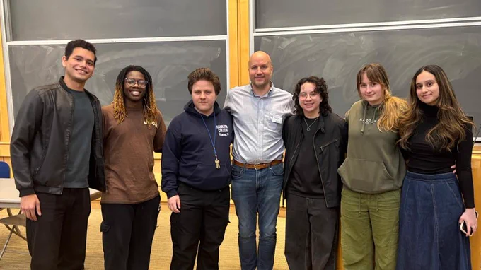 A group of seven people standing side by side in a classroom setting with large chalkboards in the background. They are casually dressed and smiling at the camera.