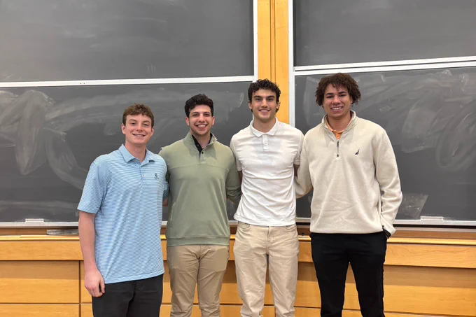 Four individuals are standing in front of a large chalkboard in a room with wooden paneling. They are all smiling and each is wearing a casual long-sleeve shirt.
