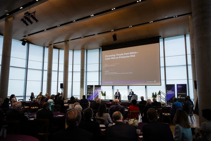 Audience attentively listening to a cybersecurity panel discussion in a modern conference room with large windows and a presentation screen.