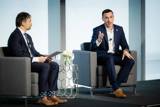 Two men in business suits engaged in a formal discussion while seated on gray armchairs on a stage.