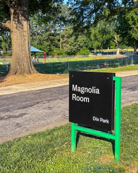 Black sign with white text reading 'Magnolia Room Dix Park' stands on green grass near a paved path and large trees.
