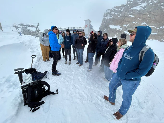 A group of students and researchers gather in the snow at high altitude near Hintertux Glacier in Austria, listening to an instructor beside scientific field equipment on a snowy, overcast day.