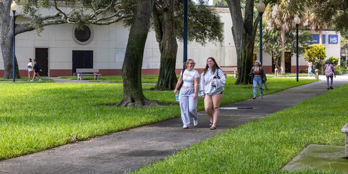 A tree-lined walkway in a grassy area with several people walking. In the background, there's a large building. Benches are scattered along the path.