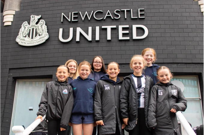 A group of young people wearing sportswear poses in front of a building with a Newcastle United sign and logo.