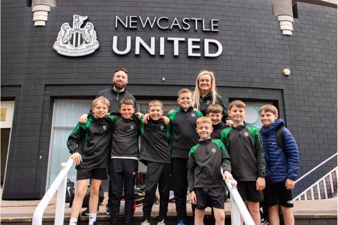 A group of people posing in front of a building with the Newcastle United sign. Eight individuals, wearing matching sports attire, stand in front of the entrance. Two adults are standing behind them, smiling.